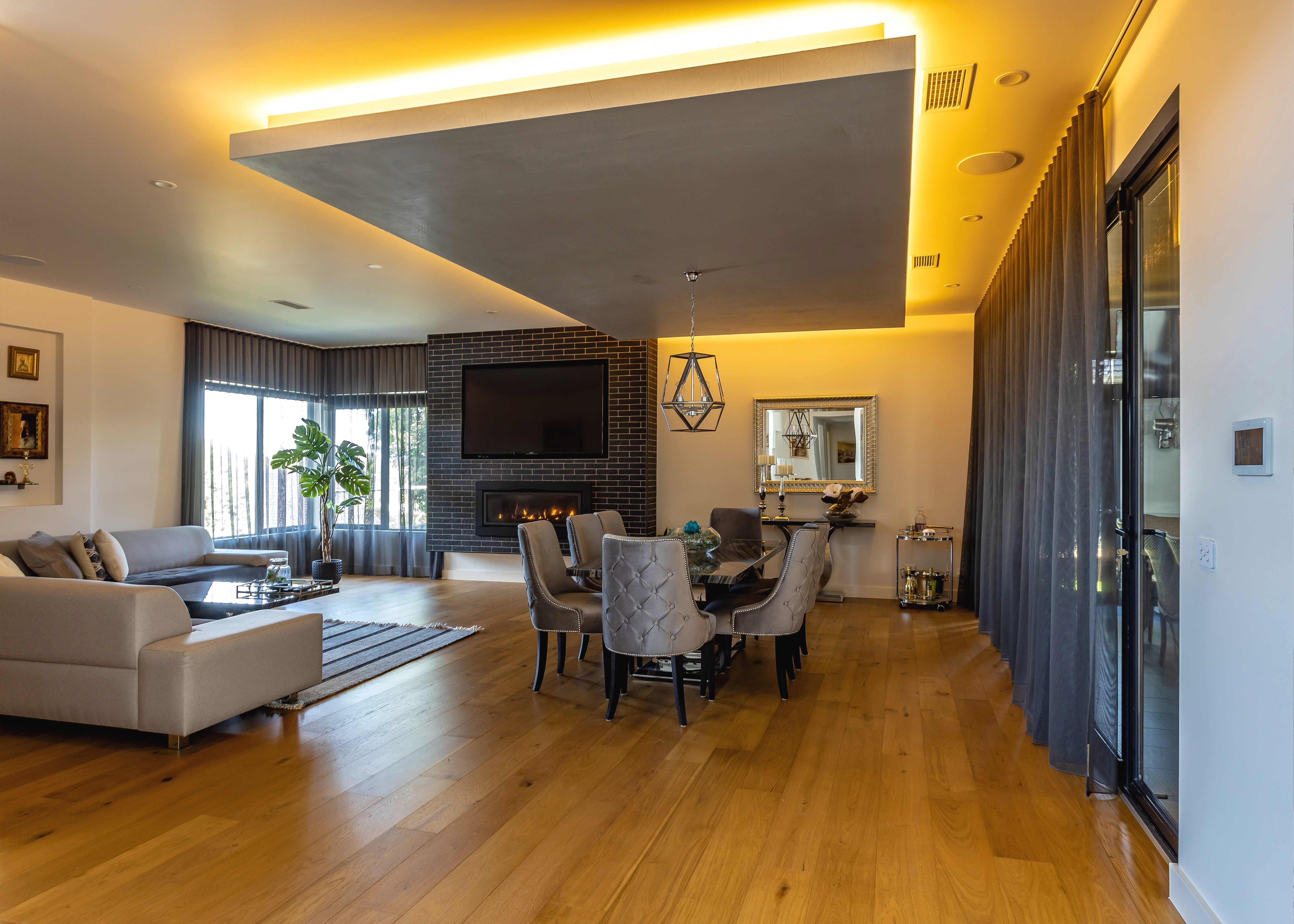 Open-plan living and dining area with timber flooring, feature ceiling detail, integrated lighting and fireplace.